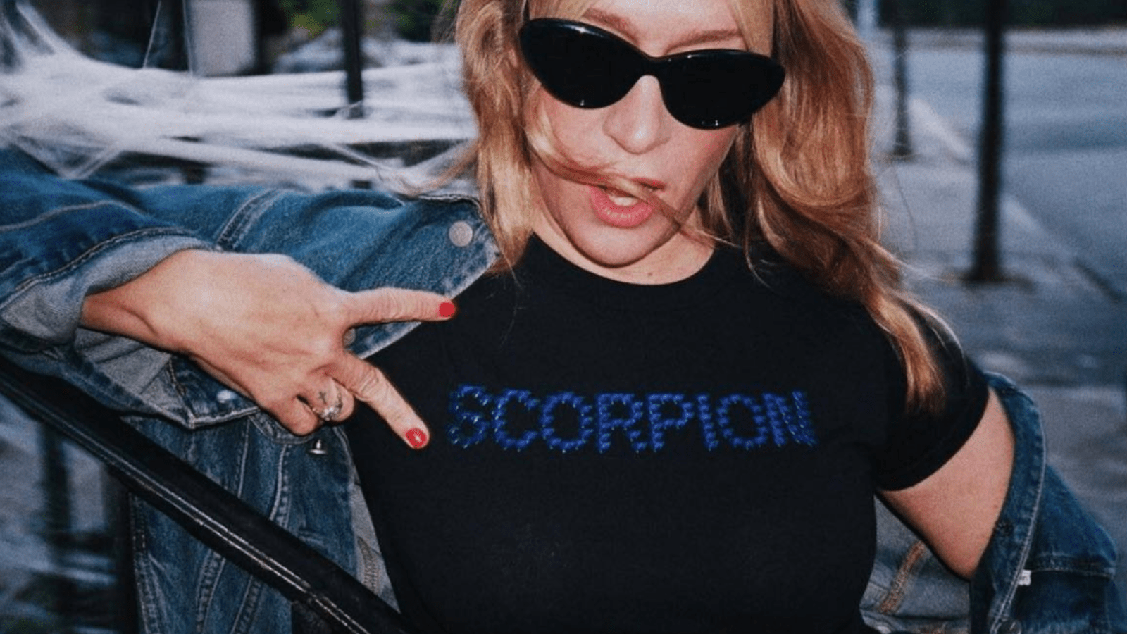 photo of blonde actress and woman chloe sevigny who is standing on a street leaning against a black metal stairwell railing. The blonde haired white woman is wearing dark black sunglasses, her hair is windswept and she is staring at the camera with right hand in a gangster style peace sign horizontally. She is wearing a short sleeved black t-shirt with the word 'scorpion' in blue jewels and she has a classic blue denim jacket slipping off her shoulders